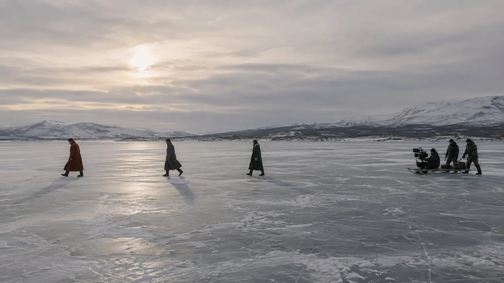 Three performers in rust, charcoal and green coats walk in a line across a vast frozen lake under a pale sun, a small camera crew on a sled to one side.