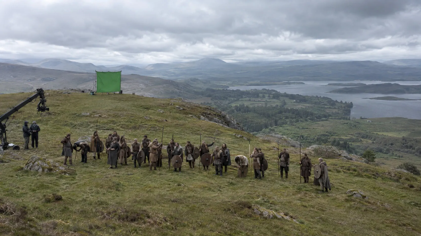 Plate: about twenty-five extras in wool tunics with spears and shields wait on a green hillside, a camera crane and a small green screen behind them.