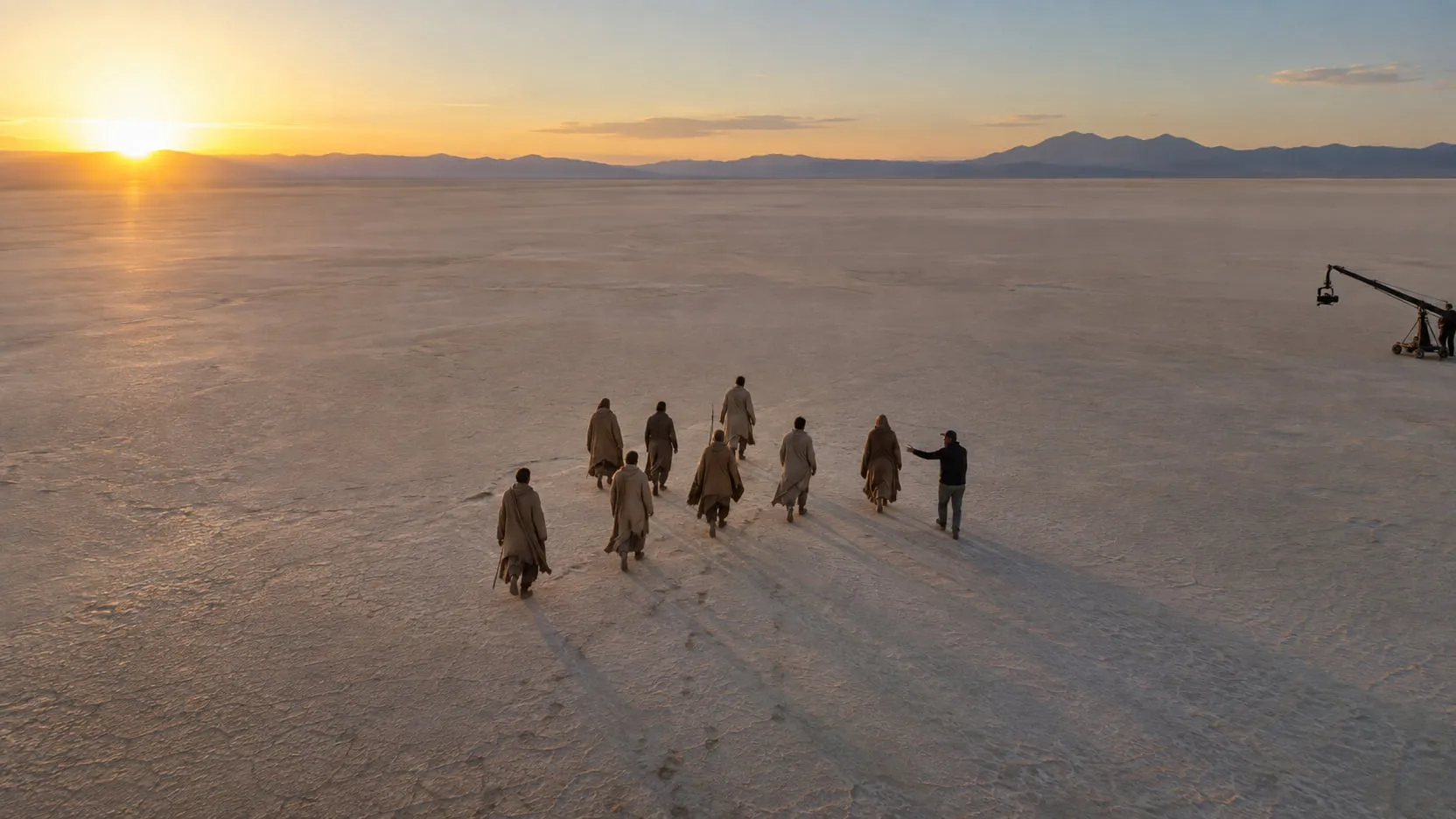 Eight extras in rough wool cloaks walk across an empty salt flat at sunset while an assistant director gestures beside them, a camera crane far to the right.