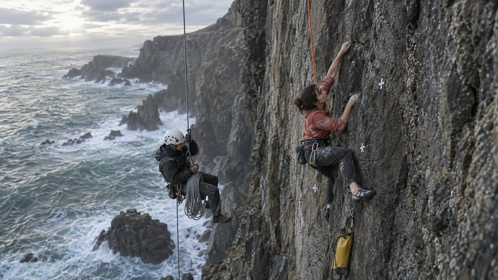 Plate: a climber hangs from a wet sea cliff above the surf, an orange safety rope running down to her, a rope-access technician beside her, tape markers on the rock and a rope bag below.