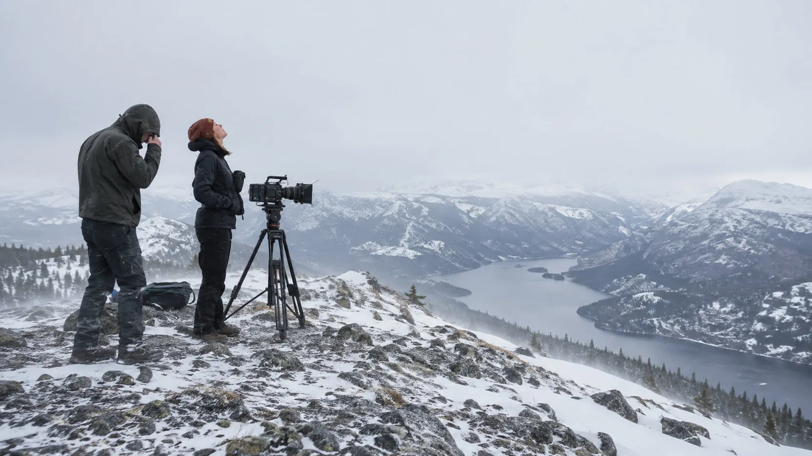 A cinematographer on a snowy ridge looks up at a flat white overcast sky beside a camera on a tripod, an assistant pulling up his hood, a lake in the valley below.