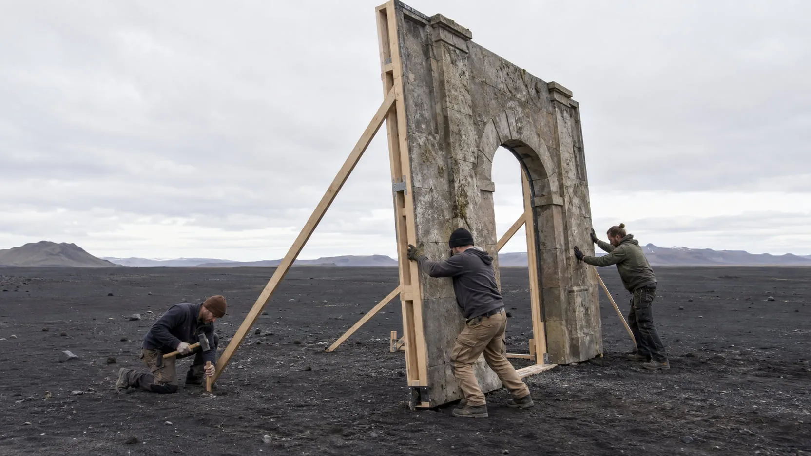 Carpenters raise and brace a partial stone-textured set wall with an arched doorway on a black volcanic plain under an overcast sky.
